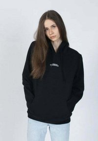 Musta huppari, jossa on naru. Vaatteessa on pieni valkoinen logo rinnassa. Yhdistetty vaaleansinisiin farkkuihin. Yksinkertainen muotoilu.