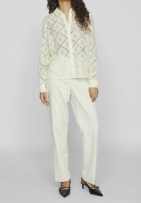 Blouse en dentelle couleur ivoire avec motif en losange, manches longues, boutonnage avant, associée à un pantalon blanc ample et des talons noirs.