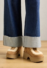 Brune suedestøvler med platform og lyse pelsfor, parret med brede denimbukser med frynset kant og en lysere denimmanchett.