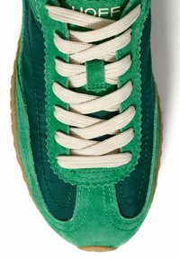 Zapatilla verde con parte superior de ante y tela texturizada, con cordones crema y suela de goma. Destaca el borde en forma de ondas a lo largo de los laterales.