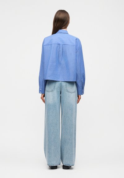 Femme vue de dos, portant une chemise à manches longues bleu rayé et un jean large bleu clair avec de petites ornements.