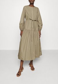 Donna che indossa un vestito midi a maniche lunghe di colore verde oliva con cintura annodata, gonna a pieghe e sandali con tacco aperti beige.