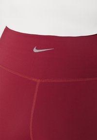 Leggings performance rossi con un elastico alto in vita, texture liscia e un logo Nike argento riflettente sulla parte anteriore. Dettaglio di doppia cucitura.