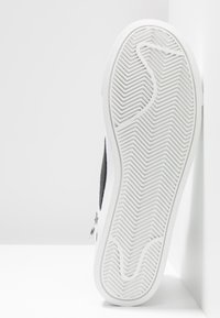 Valkoinen ja musta tennari, jossa on teksturoitu valkoinen kumipohja, jossa on siksak-kuvio ja sivuvetoketju helppoa käyttöä varten.