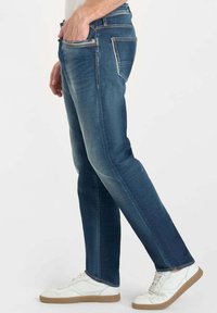 Hombre vistiendo jeans azules con una mano en el bolsillo y zapatillas blancas sobre un fondo claro.