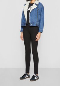 Persona que lleva una chaqueta de mezclilla forrada de sherpa azul, pantalones ajustados negros, camiseta negra con estampado blanco y zapatillas Adidas negras con franjas blancas.