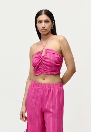 Kvinde med langt mørkt hår iført lyserød rynket halter crop top og matchende højtaljede bukser, der står mod en ensfarvet baggrund.