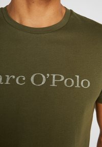 Detailní záběr na olivově zelené tričko s krátkým rukávem, na kterém je na hrudi vybledlý bílý nápis "Marc O'Polo" na horní části trupu člověka.