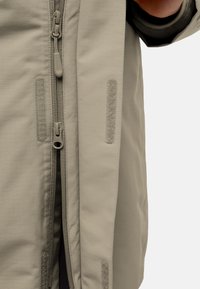Giacca beige chiaro con zip frontale, texture morbida e modelli di cuciture visibili. Include dettagli in velcro sui lati.
