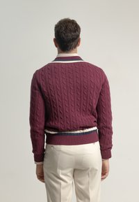 Suéter de punto en burdeos con patrón de trenzas, puños y dobladillo de canalé. Presenta un cuello a rayas en azul marino y blanco. Se lleva con pantalones de color claro.