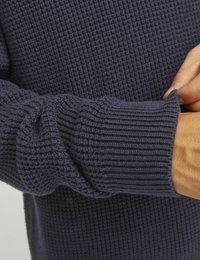 Mörkblå tröja med struktur och ribbade muddar. Närbild av ärm som visar veck i tyget och sömmens detaljer.