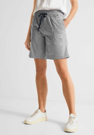 Kvinde iført grå shorts med snøre og sidelommer og hvide sneakers, stående mod en ensfarvet baggrund.