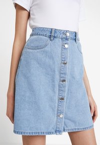 Ljusblå denimkjol med en åtsittande midja, framfickor och en rad silverknappar nedanför mitten. Avslappnad, A-linjeformat silhuett.