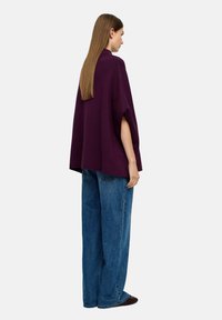 Fioletowy dziany poncho z bocznymi otworami, zestawiony z luźnymi niebieskimi dżinsami. Materiał wydaje się miękki, a design charakteryzuje się wysokim dekoltem.