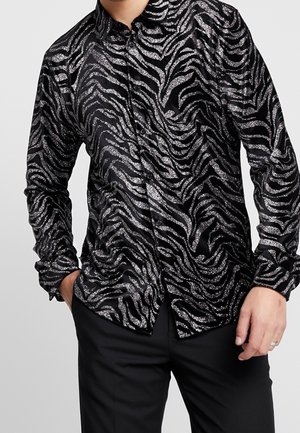 Homme portant une chemise noire à manches longues boutonnée avec des rayures zébrées argentées pailletées, pantalon noir, main gauche dans la poche, main droite relâchée sur le côté.