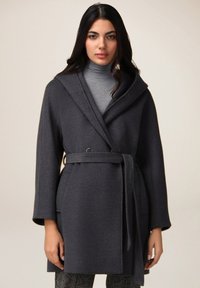Cappotto di lunghezza media grigio scuro con cappuccio, vita cintrata da una cintura e chiusura a doppio bottone. Il tessuto presenta un motivo testurizzato e dispone di tasche laterali.