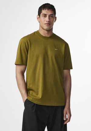Oliven grøn kortærmet t-shirt i blødt bomuld med rund hals og et lille hvidt logo på brystet, parret med sorte bukser.