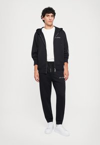 Svart zip-opp hettegenser med frontlommer og hvite detaljer, kombinert med matchende svarte joggebukser og hvite joggesko. Enkel design og avslappet passform.