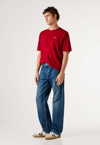 Camiseta roja de manga corta con cuello redondo, vaqueros azules de pierna recta y zapatillas beige con detalles en negro. Diseño sencillo y casual.