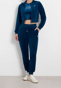 Donna che indossa una tuta in velluto blu scuro con giacca corta a zip, pantaloni coordinati con cordino e sneakers bianche, in piedi contro un muro bianco.