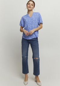 Blusa azzurro chiaro a maniche corte con scollatura a V, decorata con motivi floreali bianchi; abbinata a jeans blu strappati e scarpe beige con tacco.