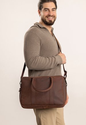 Brown leather shoulder bag with two top handles, adjustable strap, and vertical stitching. Smooth texture and minimal hardware accents.
