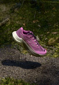 Lilla Salomon løbesko med hvid sål svævende over grus og mosdækket jord i udendørs miljø.