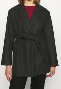 Cappotto in misto lana grigio scuro con design avvolgente, caratterizzato da una vita con cintura, rever a punta e maniche lunghe. Texture liscia e dettagli minimi.
