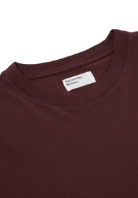 T-shirt in cotone bordeaux con scollatura rotonda; presenta un'etichetta cucita con la scritta "Universal Works". Texture liscia e design semplice.