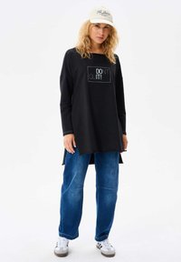 Czarna tunika z długim rękawem z białym napisem "NIE PODDAWAJ SIĘ!" z przodu, w połączeniu z luźnymi niebieskimi dżinsami i szarymi trampkami.
