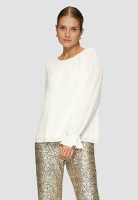 Weiße Bluse mit langen, gefältelten Ärmeln und rundem Ausschnitt. Kombiniert mit glänzenden, strukturierten goldenen Pailletten-Hosen. Einfaches, elegantes Design.