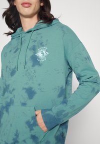 Tyrkysová mikina s kapucí a tie-dye vzorem, přední kapsa a kulatý grafický prvek s nápisem "BALANCE ENERGY" v bílé barvě. Kapuce se stahovacími šňůrkami a žebrované manžety.