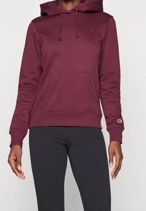 Persoon draagt een kastanjebruine hoodie met een buidelzak aan de voorkant en zwarte legging, staand tegen een effen achtergrond.