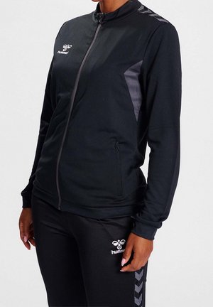 Personne portant une veste de sport noire et un pantalon avec des accents gris et un logo blanc Hummel, debout avec les mains détendues le long du corps.