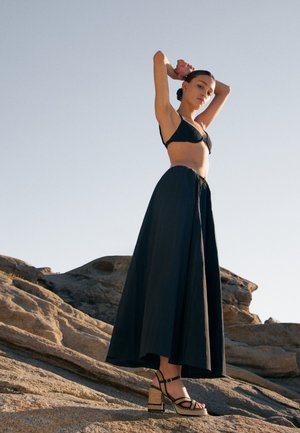 Vrouw in een zwart bikinitopje, lange zwarte rok en platformsandalen, staand op rotsachtig terrein met haar armen omhoog tegen een heldere lucht.