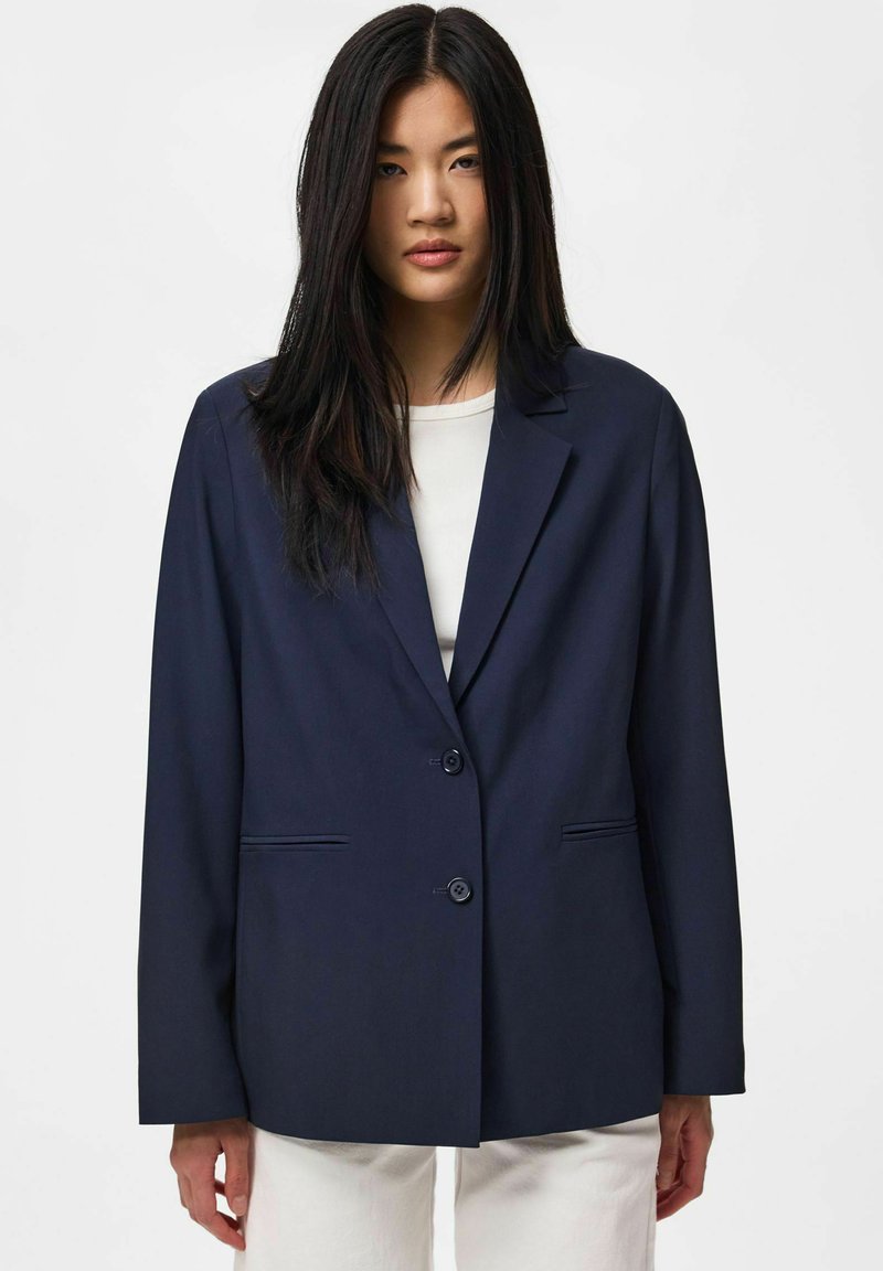 Jeune femme aux longs cheveux noirs portant un blazer bleu marine foncé sur un haut blanc et un pantalon blanc, debout devant un fond uni.