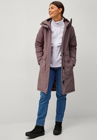 Chaqueta larga, ligera y aislante de color malva con capucha, bolsillos con cremallera y cuerdas ajustables. Se lleva sobre un forro polar gris claro con cremallera y jeans.