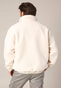 Veste polaire blanche à col haut, avec des poignets élastiques et une taille élastique. Texture douce, design simple, sans motifs ni quincaillerie visibles.