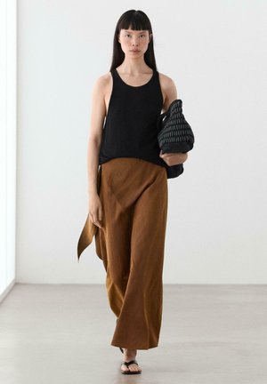 Frau mit langen dunklen Haaren, die ein ärmelloses schwarzes Top, braune Hosen mit weitem Bein, schwarze Sandalen trägt und eine strukturierte schwarze Handtasche trägt.