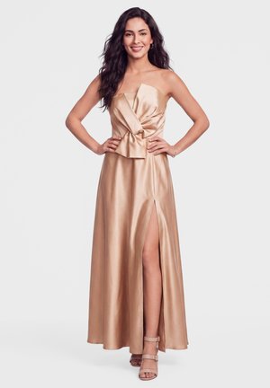 Femme debout, souriante, mains sur les hanches, portant une robe bustier beige en satin avec une fente à l'avant et des sandales à talons assorties, sur fond blanc.