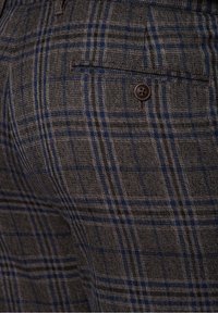 Zbliżenie na szarą i niebieską kratę materiału z pojedynczym czarnym guzikiem nad poziomą kieszenią szczelinową na ubraniu.