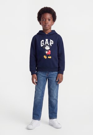 Enfant aux cheveux bouclés portant un sweat à capuche bleu marine avec le logo Mickey Mouse et GAP, un jean bleu et des baskets blanches, debout devant un fond uni.