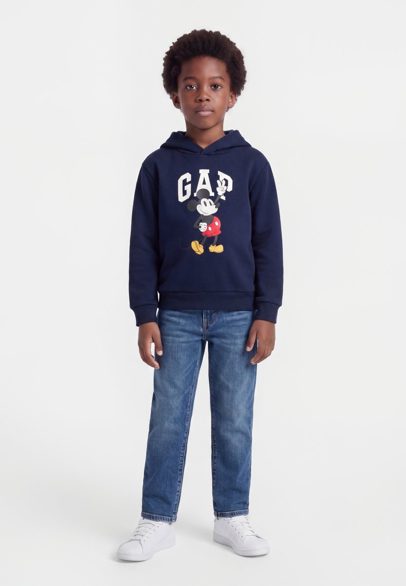 Enfant aux cheveux bouclés portant un sweat à capuche bleu marine avec le logo Mickey Mouse et GAP, un jean bleu et des baskets blanches, debout devant un fond uni.
