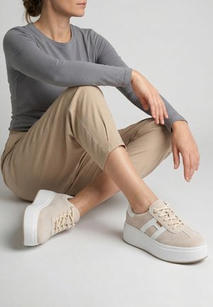 Personne en pantalon beige et haut gris à manches longues, assise en tailleur, portant des baskets compensées beiges avec semelles et lacets blancs.
