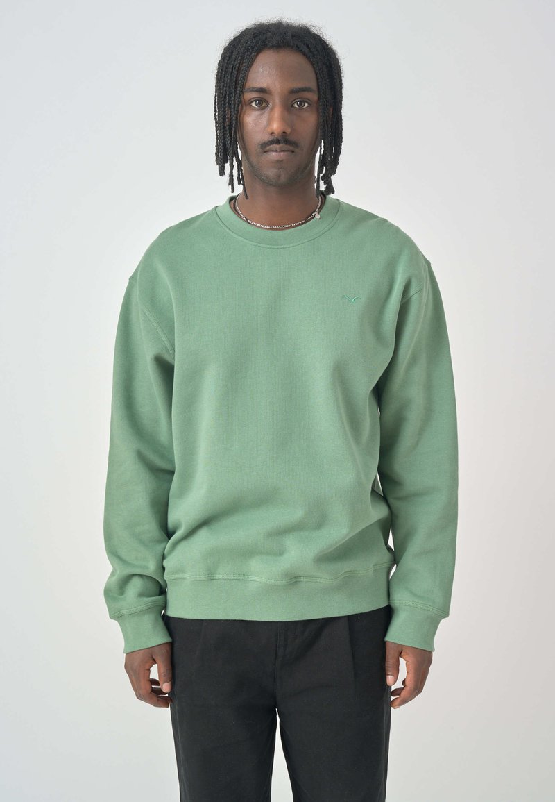 Green sweatshirt with a crew neck, wide sleeves, and ribbed cuffs. Features subtle branding in the chest area. Paired with black pants.