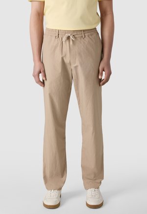 Persona che indossa pantaloni beige con coulisse, sneakers bianche e una maglietta gialla a maniche corte, in piedi contro uno sfondo uniforme.