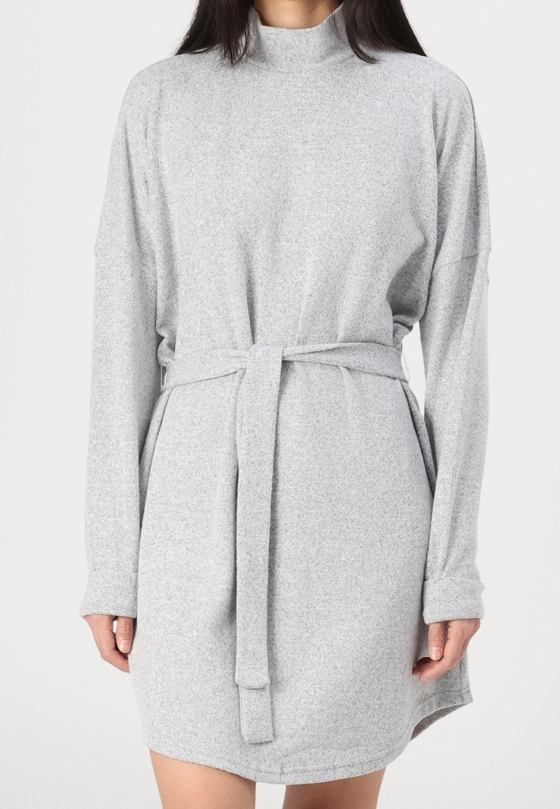 Femme portant une robe en maille gris clair à manches longues, col montant et ceinture nouée à la taille, debout devant un fond uni.