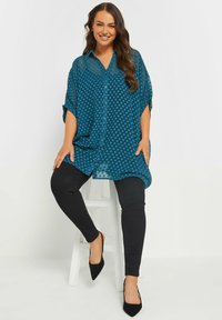 Blusa trasparente verde acqua con pois, dotata di bottoni frontali e maniche corte, abbinata a leggings neri attillati e tacchi a punta neri.