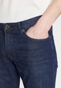 Handen vilar i den främre fickan på mörkblå denimjeans som bärs med en vit topp.