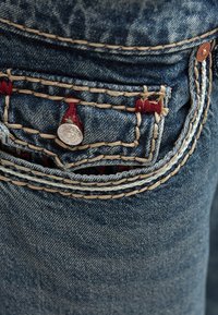 Jeansy z denimu w głębokim niebieskim kolorze, z małą przednią kieszenią ozdobioną czerwonym przeszyciem, miedzianymi dodatkami i teksturowaną tkaniną.